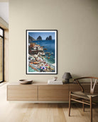 A framed vertical art print depicting a colorful coastal village and sea with rock formations, displayed on a beige wall above a wooden console table in a modern room setting.
