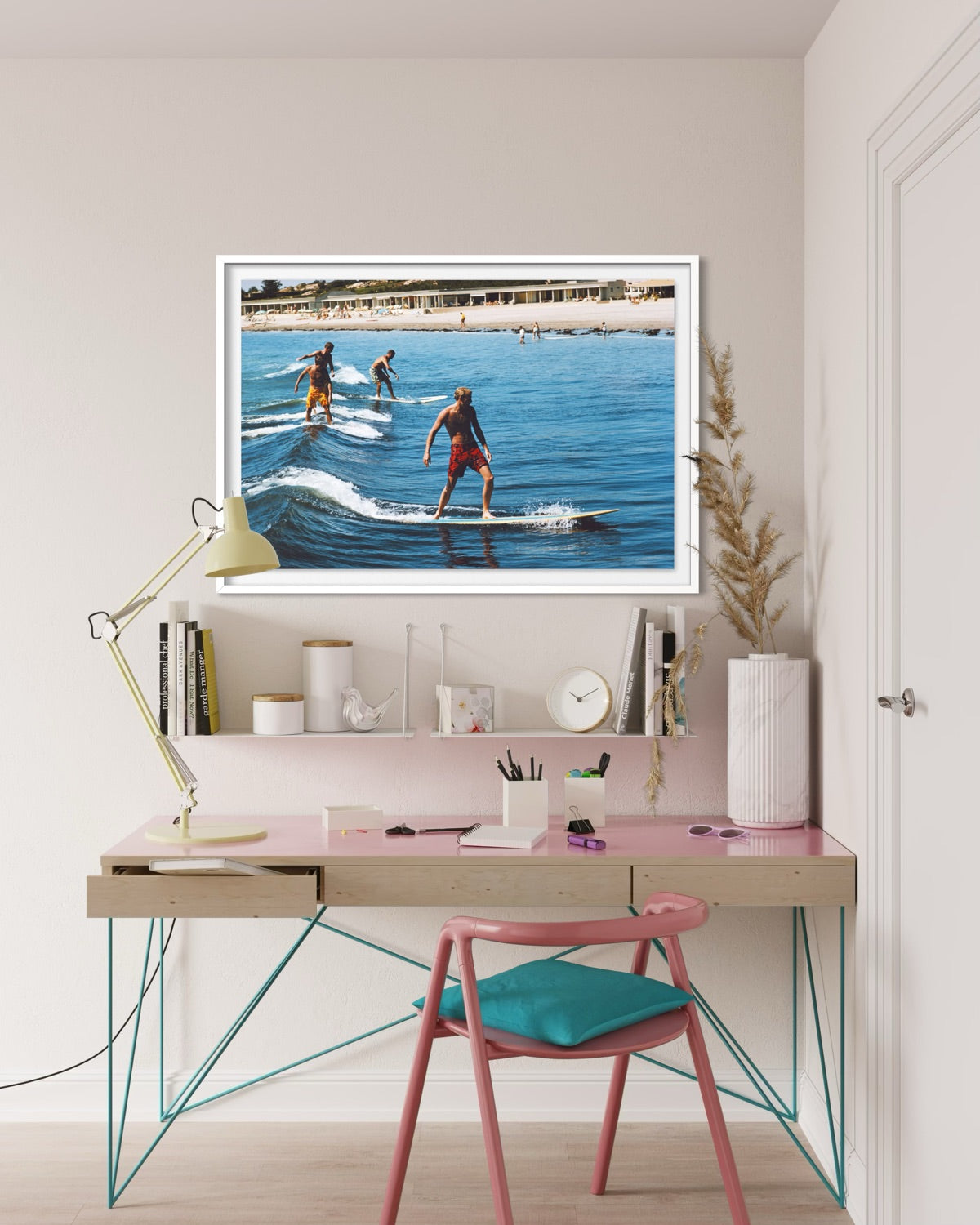 A framed art print depicting three surfers riding waves in a sunny beach setting, displayed on a light pink desk shelf in a modern, styled home office environment with a desk, chair, and lamp.