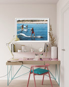 A framed art print depicting three surfers riding waves in a sunny beach setting, displayed on a light pink desk shelf in a modern, styled home office environment with a desk, chair, and lamp.
