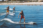 A man in red patterned swim trunks stands on a surfboard in blue ocean water, with other surfers and a beach with buildings in the background.