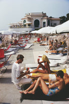 A sunlit resort poolside scene with people lounging on white lounge chairs under large umbrellas, a waiter serving drinks, and a large white building in the background.