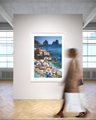 A framed coastal art print depicting a colorful seaside village with boats and rocky cliffs, displayed on a wall in a modern gallery space with a blurred person walking past in the foreground.