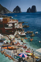 A vibrant coastal beach scene with people swimming and relaxing in clear blue water, boats on the shore, a red building with outdoor seating, and large rock formations rising from the sea under a blue sky.