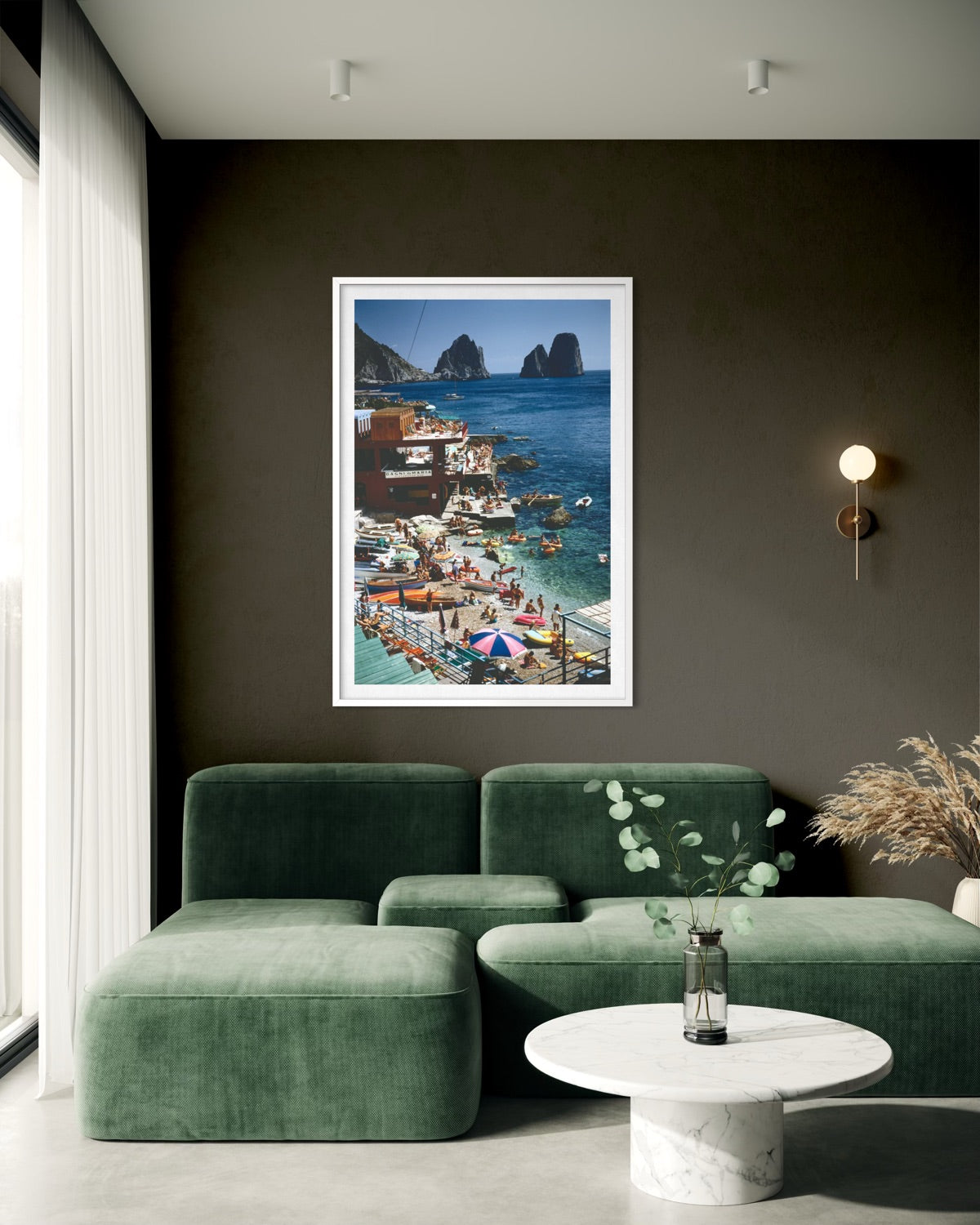 A framed art print of a colorful coastal village and harbor scene with boats and blue water, displayed on a dark wall above a green sectional sofa in a modern living room.