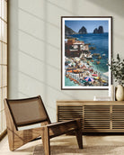A framed vertical art print depicting a vibrant coastal harbor scene with boats, colorful umbrellas, and rocky cliffs, styled in a modern interior next to a wooden chair and console table.