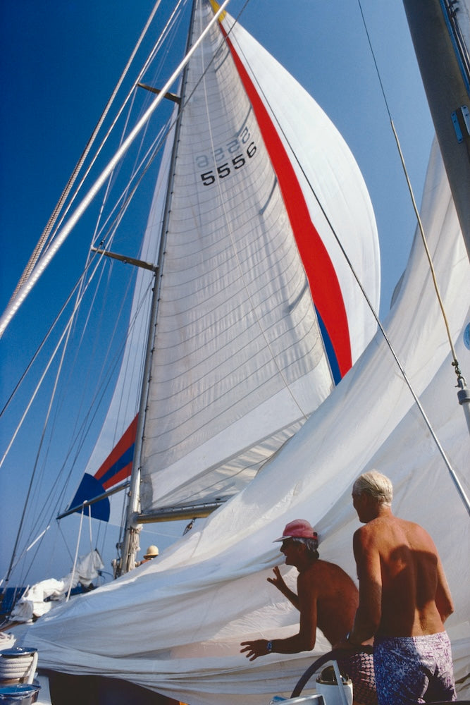 A close-up action shot of a sailboat under a bright blue sky, showing crew members handling the large white mainsail with red and blue stripes. The number 5556 is printed on the sail.