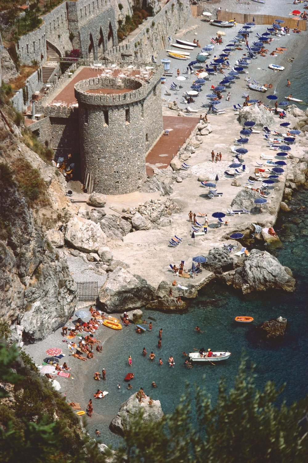 A high-angle view of a rocky coastal beach and harbor with a stone tower, many blue umbrellas, small boats, swimmers, and people along the shoreline.