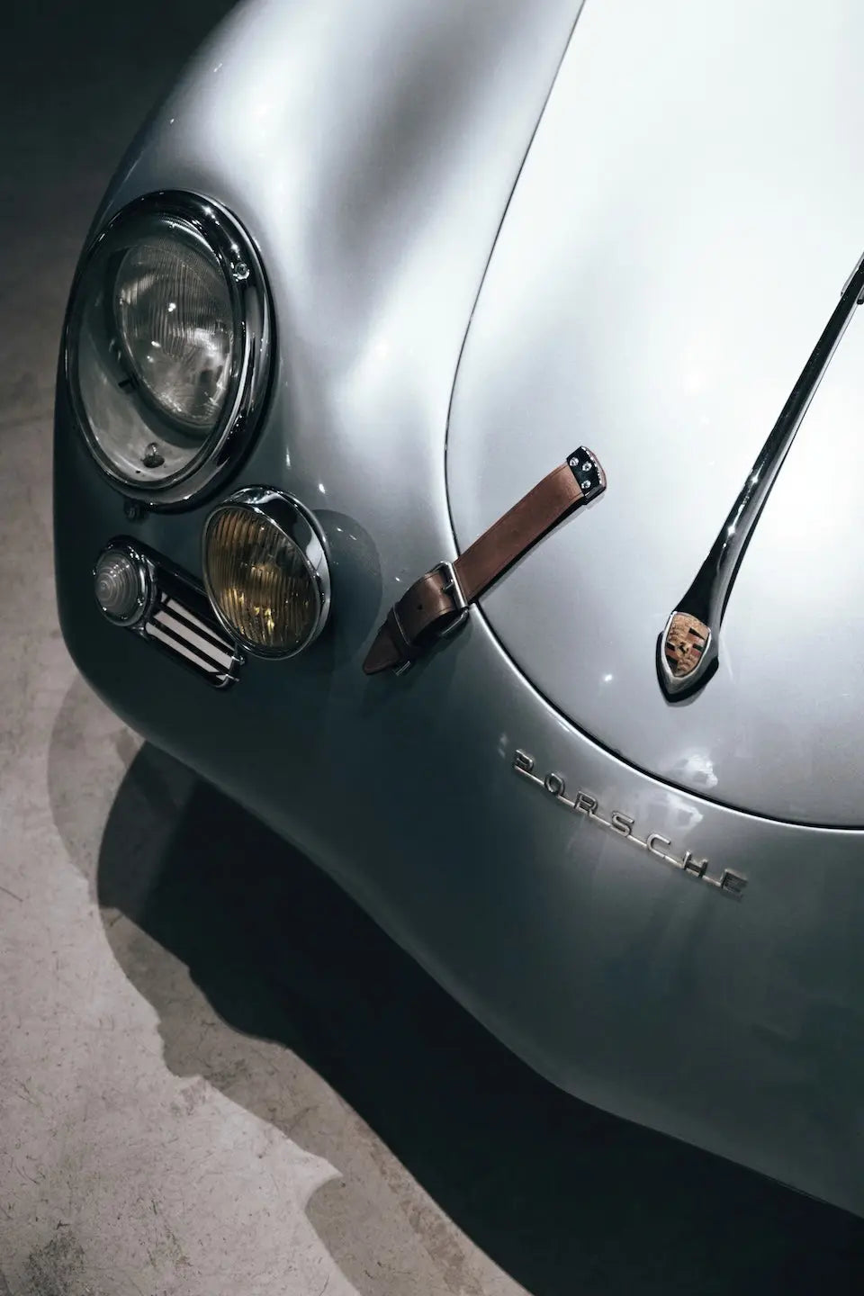 Close-up of a silver Porsche car body with the Porsche crest badge, headlight, and a brown leather wristlet strap resting on the hood.