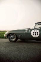 A dark green classic sports car photographed from a low side angle on an asphalt road with a pale sky and grass in the background. The side shows a large white racing number 111 decal with EFG and Columbia branding.