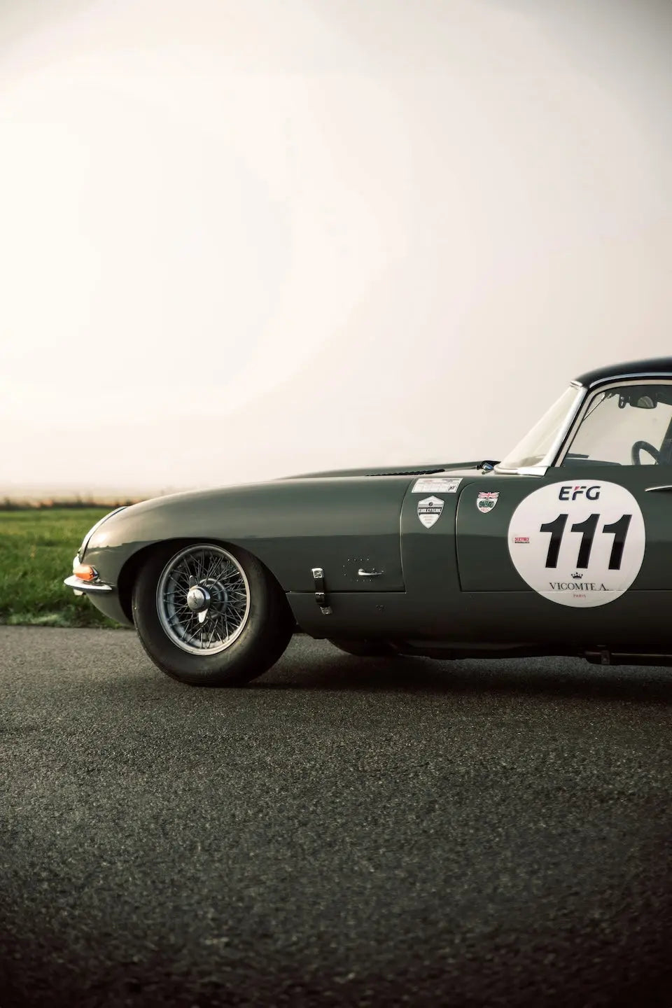 A dark green classic sports car photographed from a low side angle on an asphalt road with a pale sky and grass in the background. The side shows a large white racing number 111 decal with EFG and Columbia branding.
