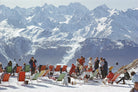 A snowy mountain landscape with many people sitting and standing in colorful deck chairs on the snow, with large snow-covered peaks in the background. The Picturalist Wall Art and Photography for Designed Interiors. Slim Aarons Collection, Skiing. Getty Images Gallery. 