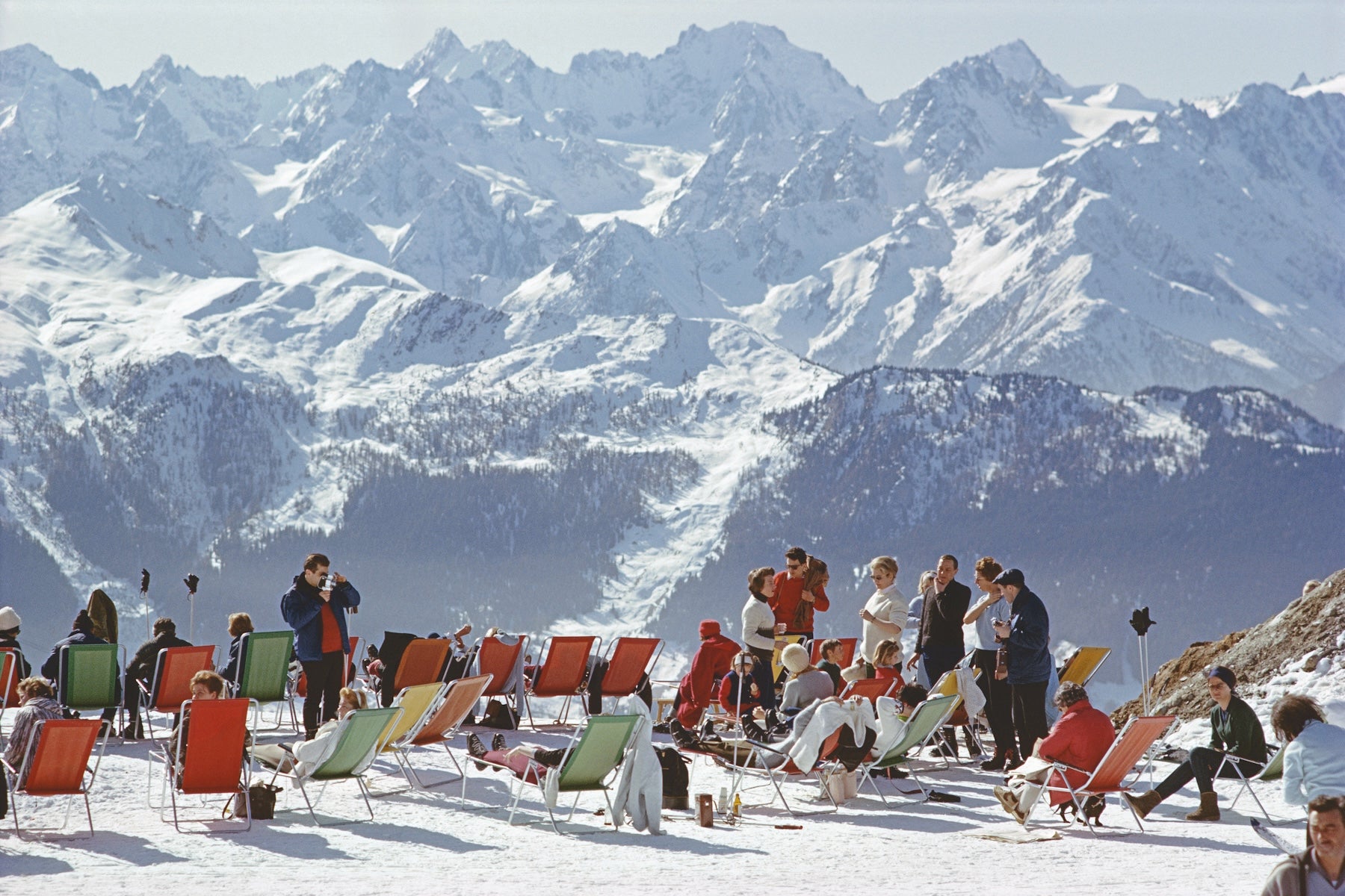 A snowy mountain landscape with many people sitting and standing in colorful deck chairs on the snow, with large snow-covered peaks in the background. The Picturalist Wall Art and Photography for Designed Interiors. Slim Aarons Collection, Skiing. Getty Images Gallery. 