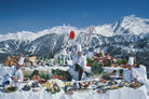 A large outdoor food spread arranged on snow in a mountain setting, with chefs in white uniforms standing behind it, plates of food, bottles, and a red flag against a blue sky and snowy peaks.