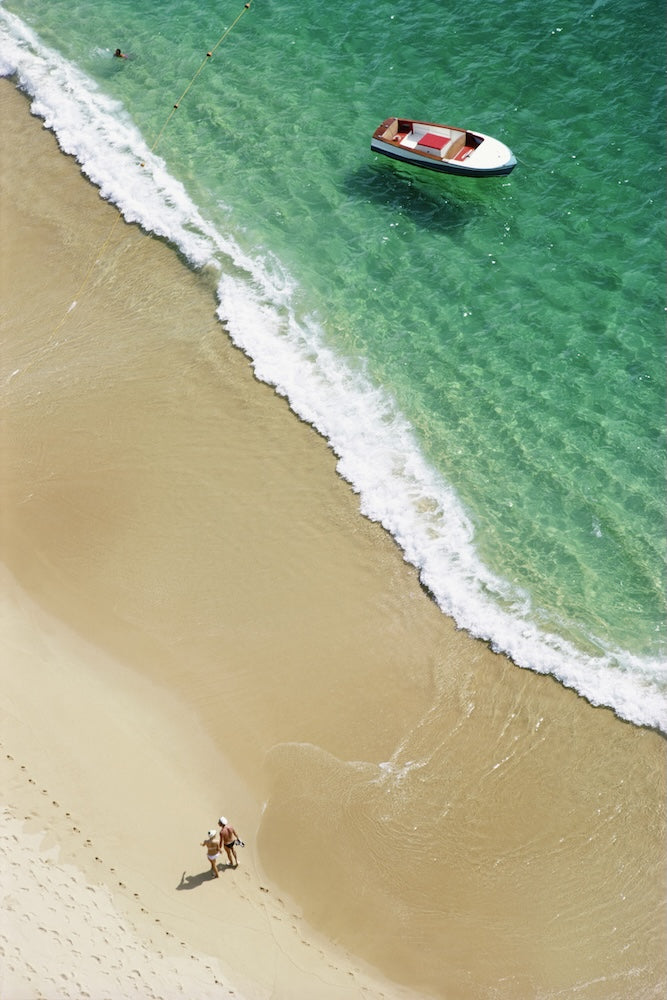 Aerial beach scene with turquoise ocean water, white surf, pale sand, a small white boat anchored in the water, and two people walking near the shoreline.
