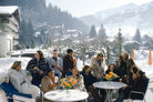 A group of people sitting and standing around small outdoor tables in a snowy mountain village setting, wearing winter clothing and holding drinks. Snow-covered trees, chalets, and mountains are visible in the background.  The Picturalist Wall Art and Photography for Designed Interiors. Slim Aarons Collection, Skiing. Getty Images Gallery. 