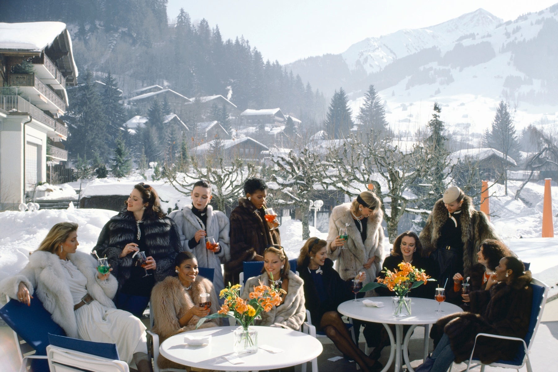 A group of people sitting and standing around small outdoor tables in a snowy mountain village setting, wearing winter clothing and holding drinks. Snow-covered trees, chalets, and mountains are visible in the background.  The Picturalist Wall Art and Photography for Designed Interiors. Slim Aarons Collection, Skiing. Getty Images Gallery. 
