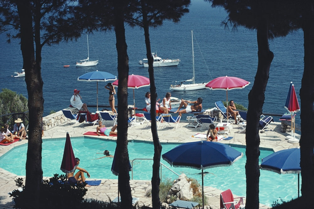 A scenic coastal resort scene with a swimming pool, lounge chairs, colorful umbrellas, people relaxing, sailboats on the blue sea, and dark tree trunks framing the foreground.  The Picturalist Wall Art and Photography for Designed Interiors. Slim Aarons Collection, Poolside. Getty Images Gallery. 