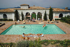 A large outdoor swimming pool with several people relaxing in and around it, set in front of a white stone villa with terracotta roofs. Sun loungers, umbrellas, and tiled decking surround the pool. The Picturalist Wall Art and Photography for Designed Interiors. Slim Aarons Collection, Poolside. Getty Images Gallery. 