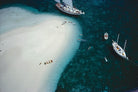 Aerial view of a tropical sandbar with clear turquoise water, several sailboats anchored nearby, and small swimmers in the ocean. The Picturalist Wall Art and Photography for Designed Interiors. 
