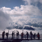 A scenic mountain landscape with snow-covered peaks rising above a sea of clouds, and a group of silhouetted people standing along a wooden railing holding skis in a high-altitude alpine setting.