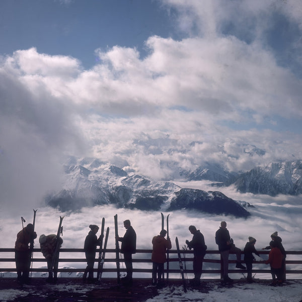 A scenic mountain landscape with snow-covered peaks rising above a sea of clouds, and a group of silhouetted people standing along a wooden railing holding skis in a high-altitude alpine setting.