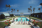 Two women and two children standing by a pool with palm trees and a clear blue sky.  The Picturalist Slim Aarons Collection
