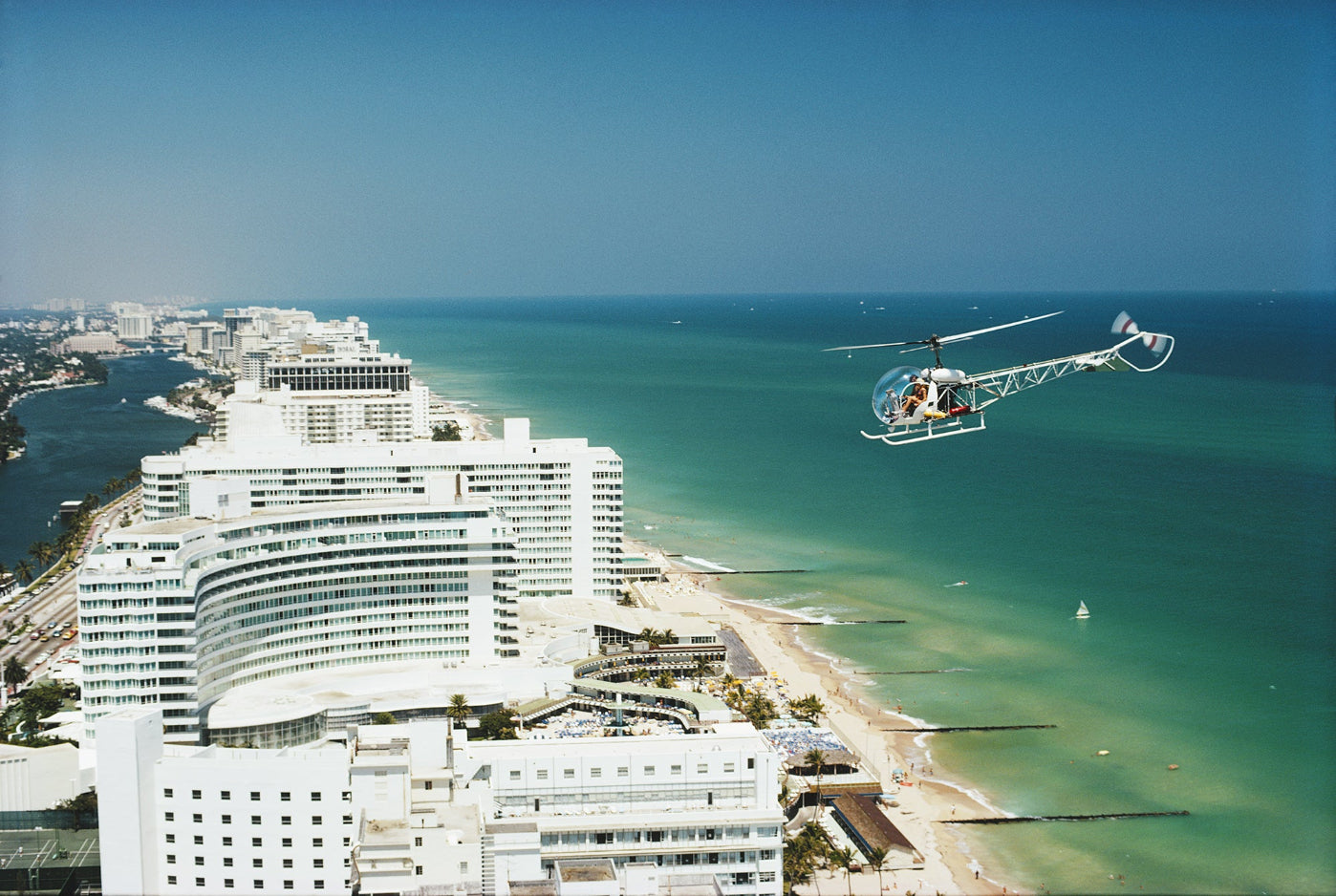 Helicopter flying over a coastal city with buildings and ocean.  The Picturalist Slim Aarons Collection