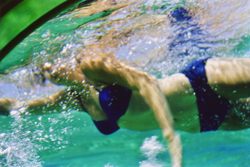 A person swimming underwater in bright turquoise pool water, wearing dark blue swim trunks and a dark blue swim cap.  The Picturalist Wall Art and Photography for Designed Interiors. Slim Aarons Collection, Poolside. Getty Images Gallery. 