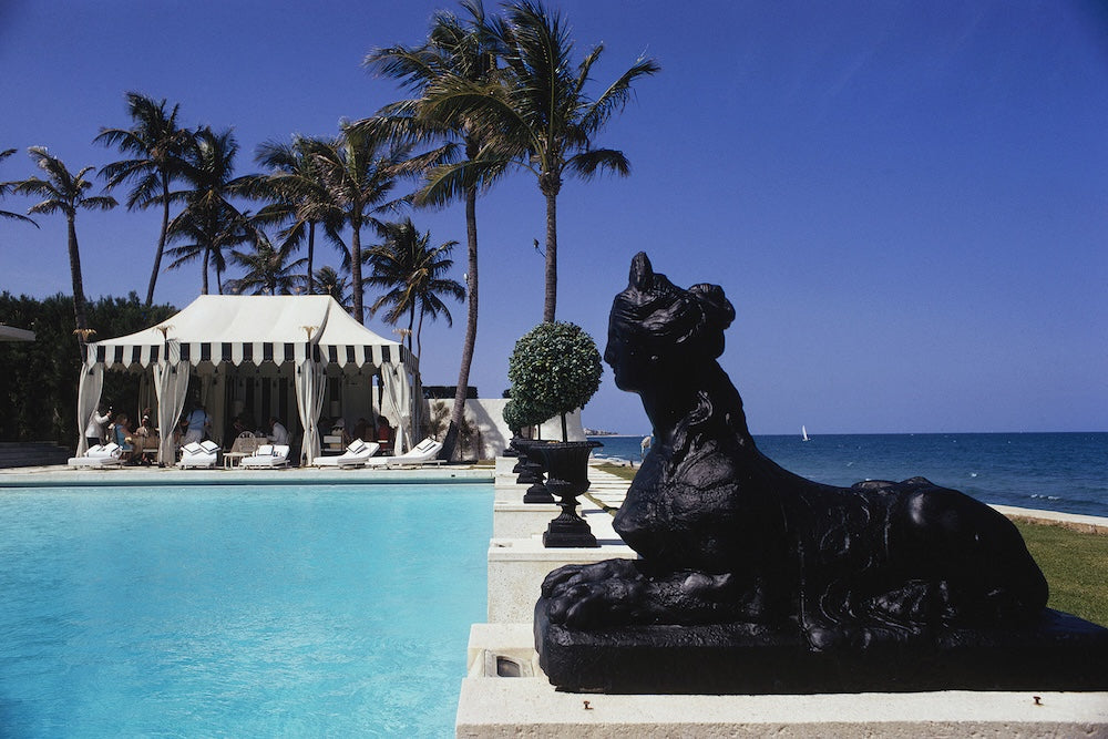 A luxurious resort scene with a bright blue swimming pool in the foreground, a black lion statue, white cabanas, tall palm trees, and the blue ocean under a clear sky.  The Picturalist Slim Aarons Collection. 