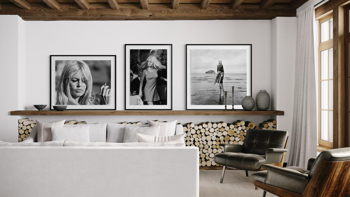 Three framed black and white art prints of French Actress Brigitte Bardot are displayed on a white wall above a wooden shelf in a modern, rustic living room setting with a white sofa, leather armchair, and wood ceiling beams.