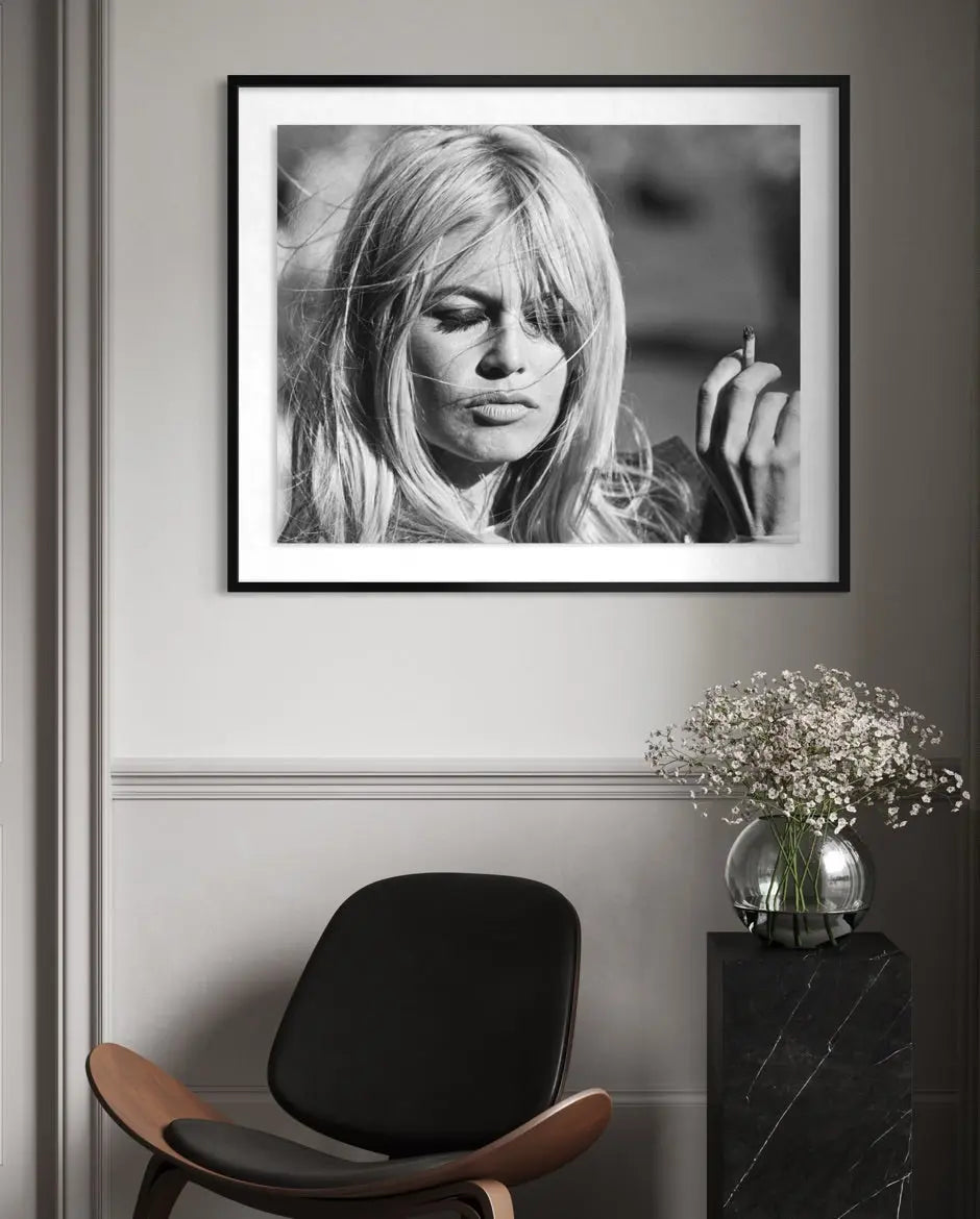 A framed black and white portrait print of actress Brigitte Bardot hanging on a gray wall above a modern chair and a vase of flowers.