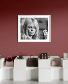 A framed black and white portrait print of actress Brigitte Bardot with long blonde hair, displayed on a dark red wall above a white display shelf with handbags.