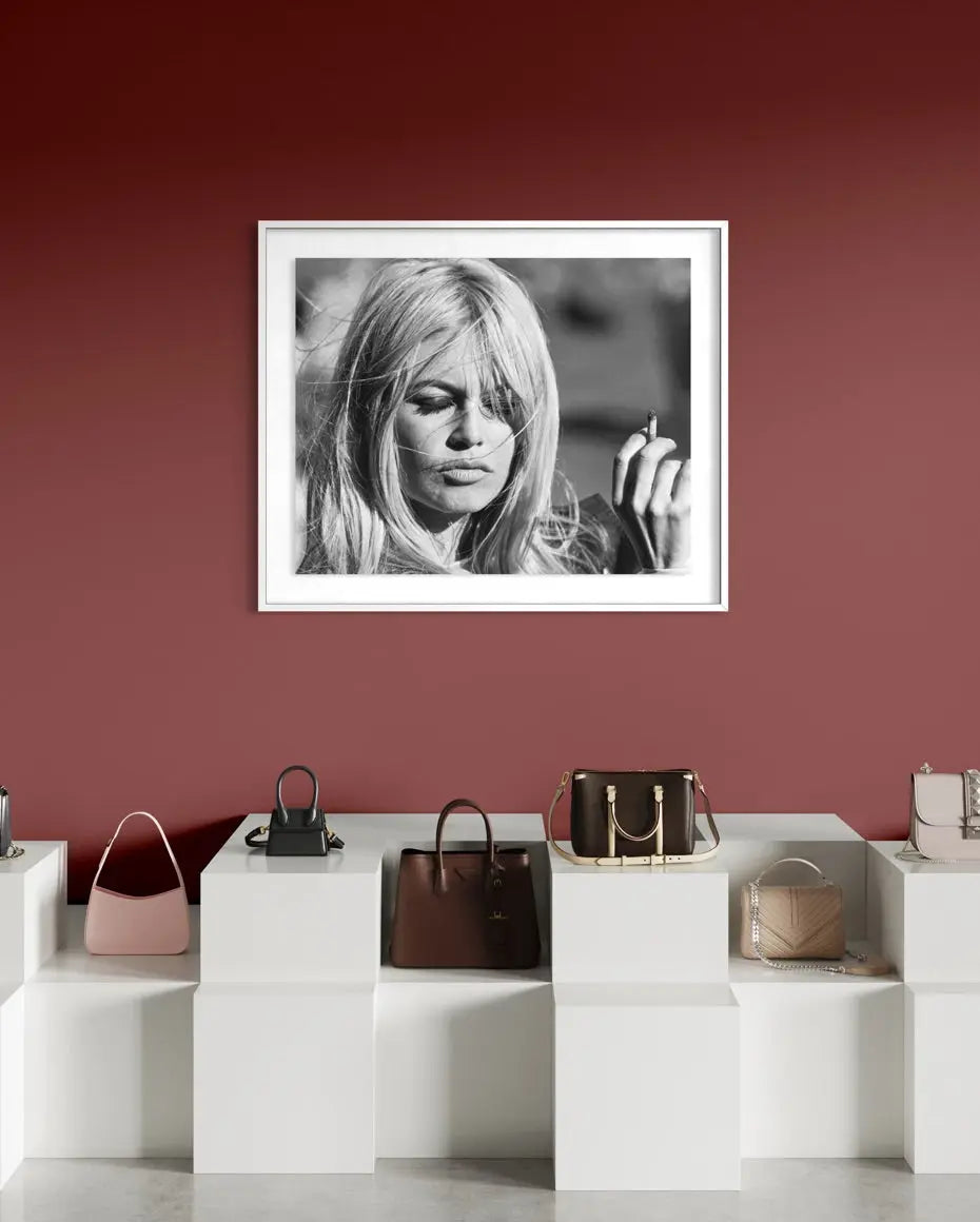 A framed black and white portrait print of actress Brigitte Bardot with long blonde hair, displayed on a dark red wall above a white display shelf with handbags.