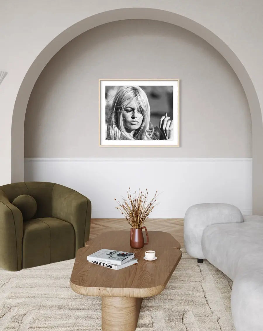 A black and white framed portrait print of actress Brigitte Bardot hanging on a beige wall in a modern living room setting with a wooden coffee table, green armchair, and light gray sofa.