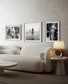Three framed black and white portrait and beach photographs hang on a textured light gray wall above a cream boucle sofa in a modern living room setting.