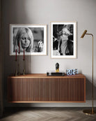Two framed black and white art prints of iconic blonde women are displayed on a light gray wall above a modern wooden sideboard. The room is styled with a brass floor lamp, decorative candlesticks, and a small bust.