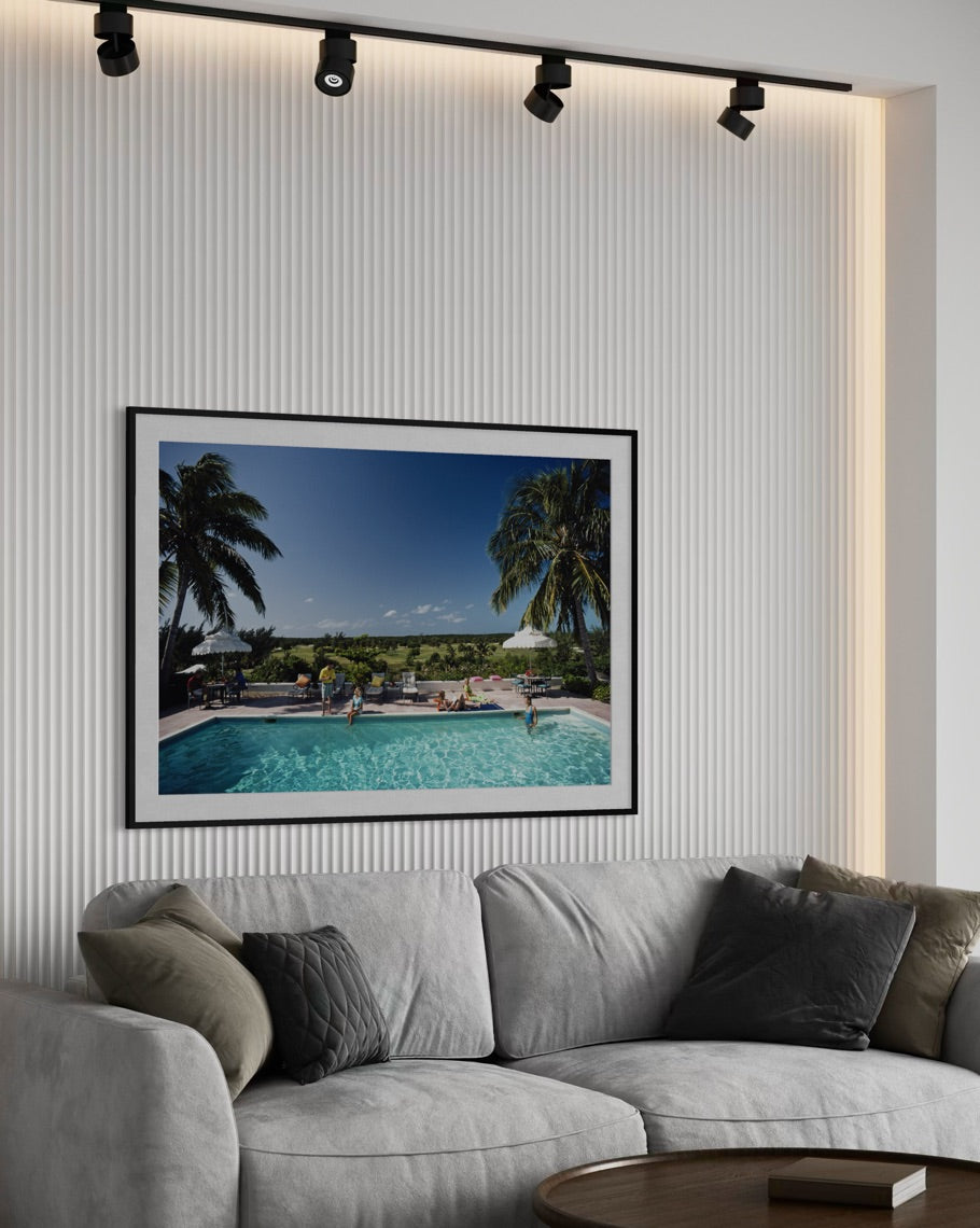 A framed art print depicting a tropical resort pool scene with palm trees and a blue sky, displayed on a ribbed white wall above a modern gray sofa in a living room setting.