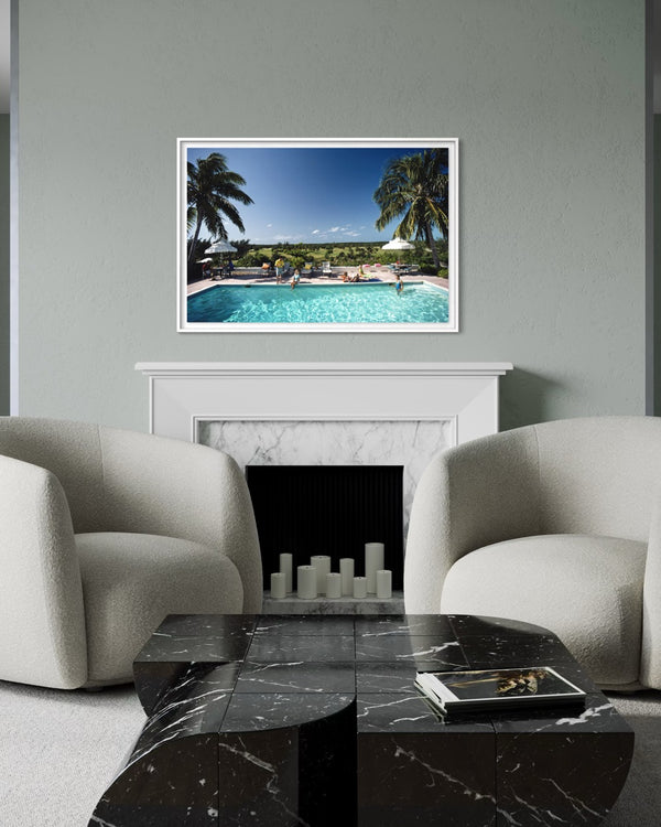 A framed art print depicting a tropical poolside scene with palm trees and a blue sky, displayed on a gray wall above a white fireplace mantel in a modern living room setting with two cream armchairs and a black marble coffee table.
