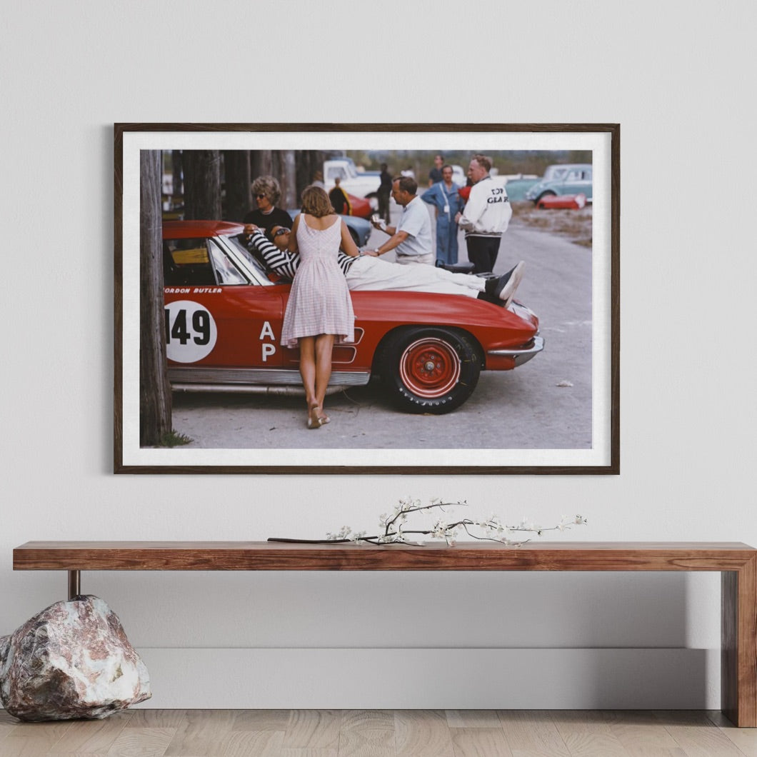 A framed vintage photograph print hanging on a light gray wall above a wooden console table. The print shows a red race car with several people standing beside it, and the frame is mounted on the wall.