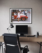 A framed art print depicting a vintage red race car scene with a woman in a white dress, displayed on a textured gray wall above a modern desk setup with a computer monitor, black office chair, and desk lamp.