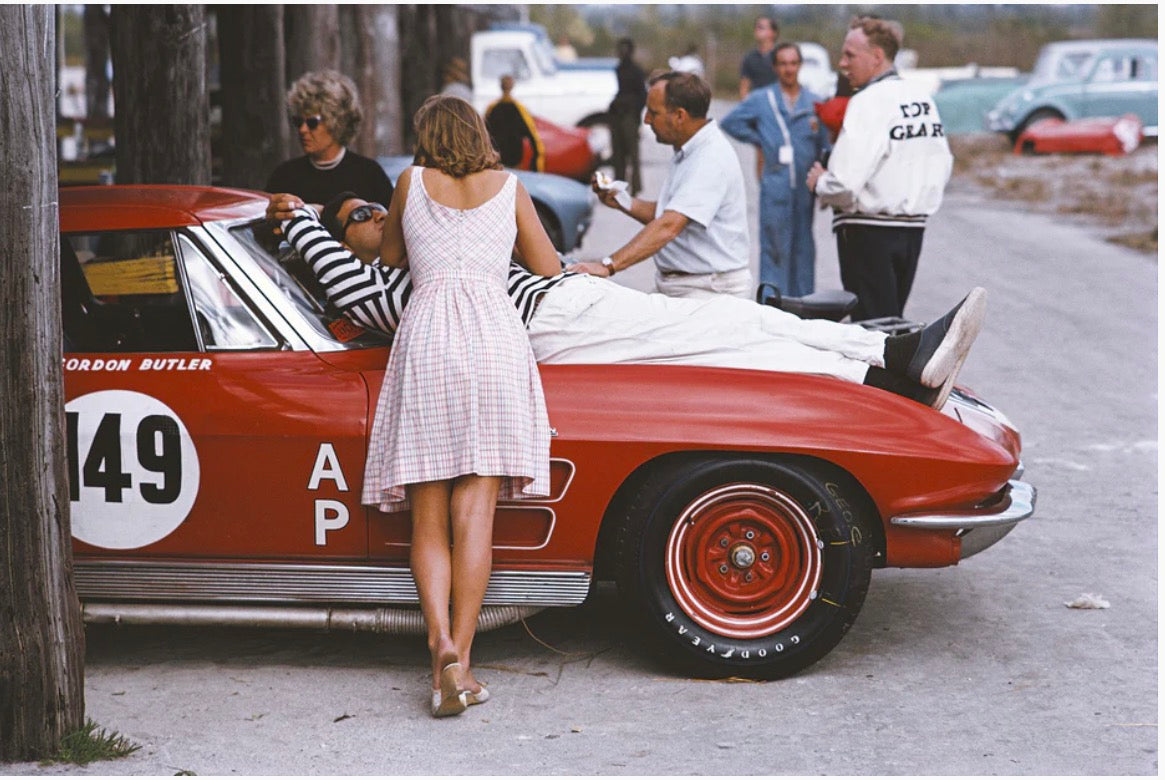 A vintage red race car with white racing decals and a driver lying on a stretcher outside, with several people standing around it in a real outdoor setting.