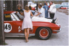 A vintage red race car with white racing decals and a driver lying on a stretcher outside, with several people standing around it in a real outdoor setting.