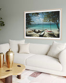 A framed beach scene print hangs on a light gray wall above a white sofa in a styled living room. The artwork shows a tropical shoreline with palm trees, blue water, and small boats, while the room background includes a white couch, pillows, a wooden coffee table, and gold vases.