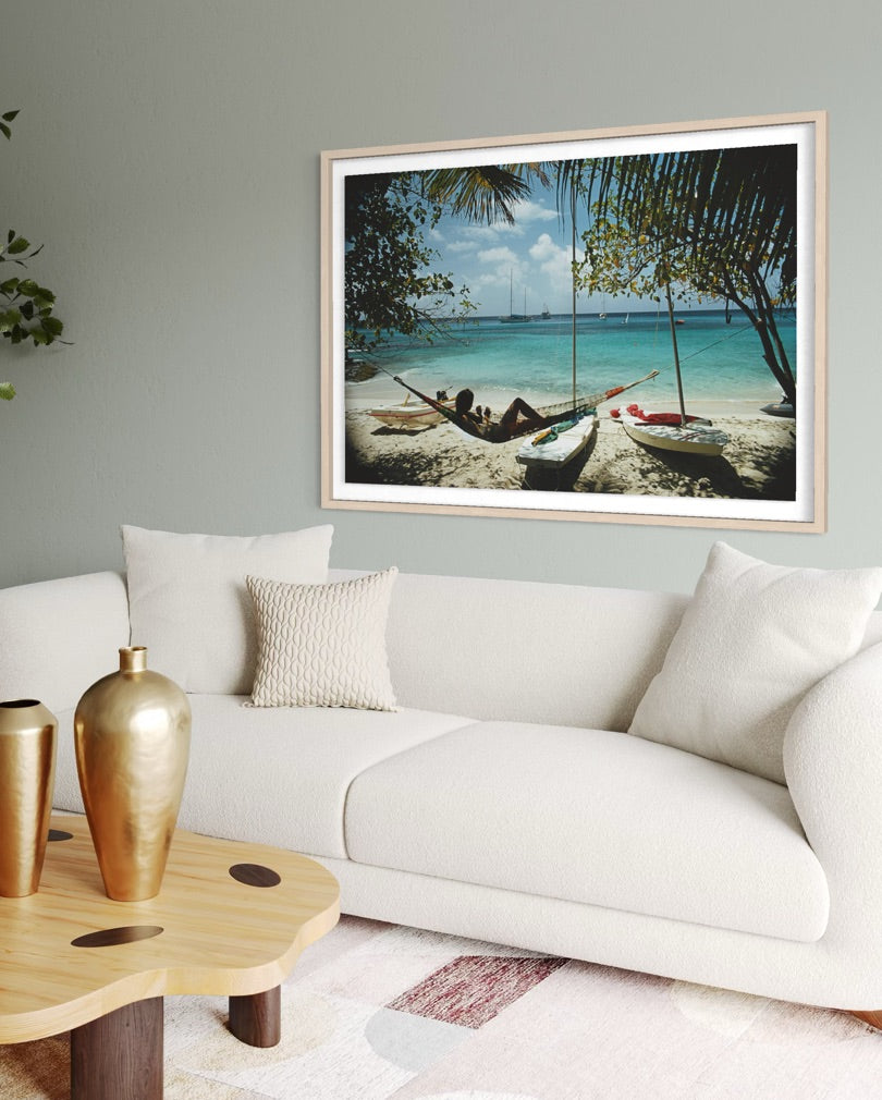 A framed beach scene print hangs on a light gray wall above a white sofa in a styled living room. The artwork shows a tropical shoreline with palm trees, blue water, and small boats, while the room background includes a white couch, pillows, a wooden coffee table, and gold vases.