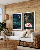 Two vertical framed art prints depicting tropical beach and pool scenes, displayed side-by-side on a rustic wood-paneled wall above a white and wicker sofa in a modern living room setting.