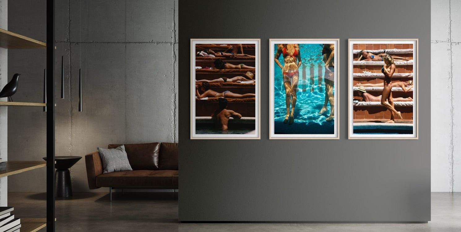 A three-panel framed wall art set hangs on a dark gray wall in a modern living room. The artwork depicts sunbathing figures on wooden decks and a swimmer in bright blue water, each panel in a white frame.