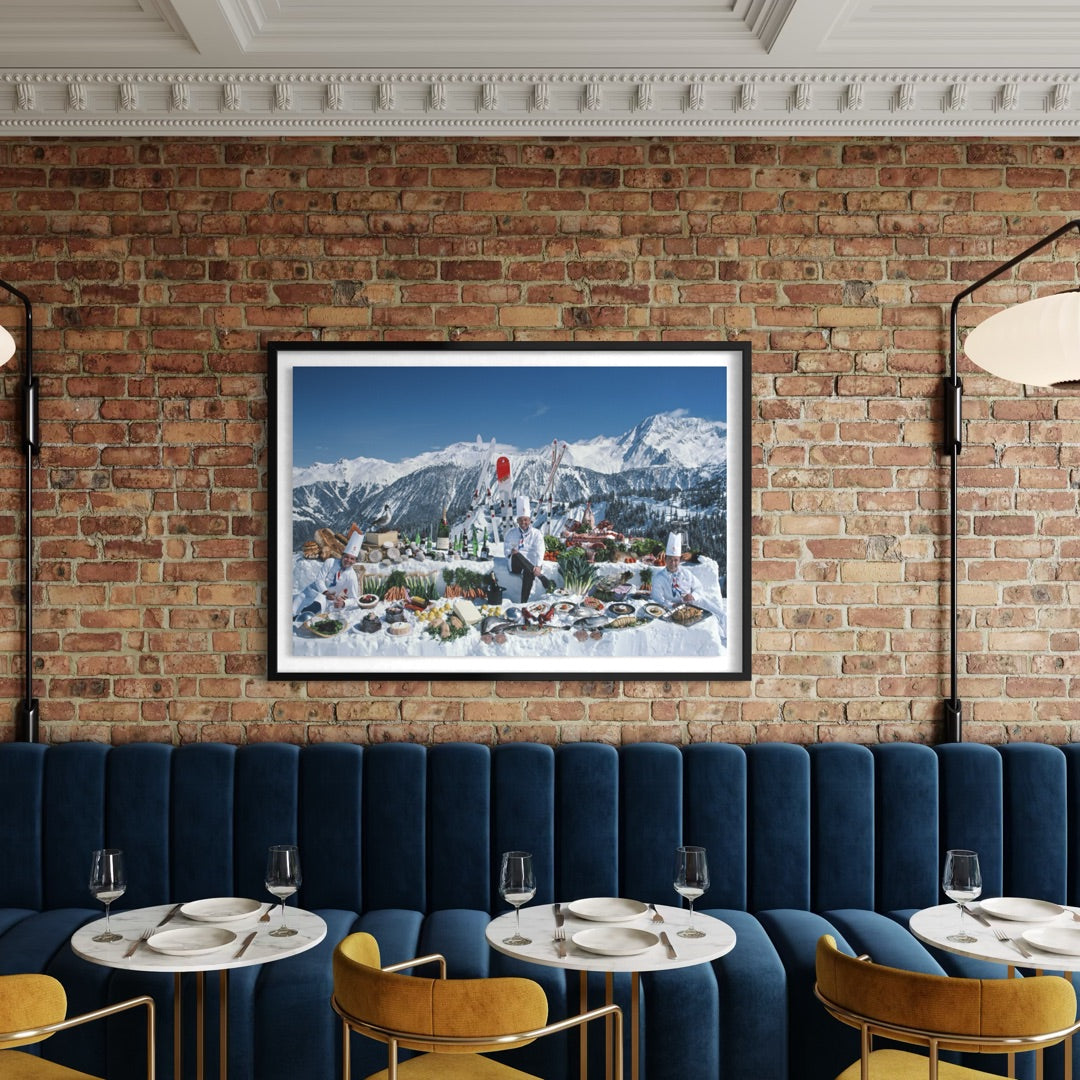 A framed art print depicting a snowy mountain village with a person holding a red balloon, displayed on a brick wall above a blue velvet booth in a restaurant setting.