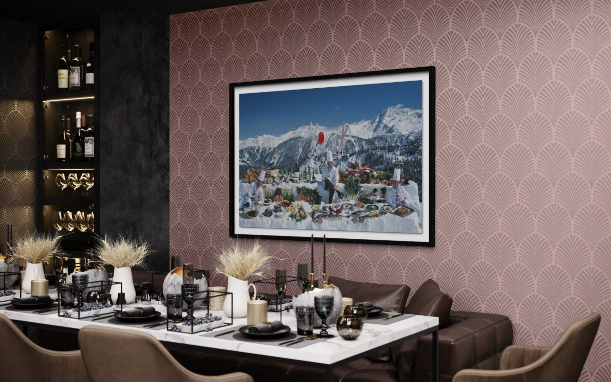 A framed art print depicting a snowy mountain town under a blue sky, displayed on a patterned pink wall above a styled dining table in a modern dining room setting.