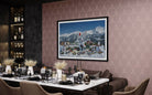 A framed art print depicting a snowy mountain town under a blue sky, displayed on a patterned pink wall above a styled dining table in a modern dining room setting.