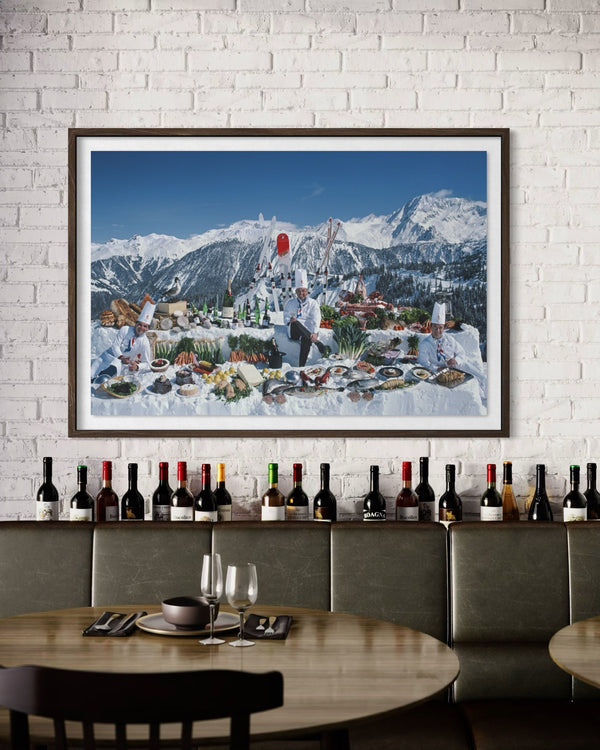 A framed art print depicting a winter mountain village with chefs preparing food, displayed on a white brick wall above a restaurant counter with wine bottles and a wooden table.